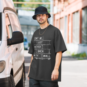 Man in bucket hat en ketting draagt oversized statement t-shirt van 100% katoen met tekst "Don't be just another brick in the wall" in antraciet met witte bakstenen print, stedelijke achtergrond met bestelwagen.