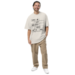 Man draagt oversized statement t-shirt van 100% katoen met tekst "Don't be just another brick in the wall" in zandkleur met bakstenen muur print, gecombineerd met kaki cargobroek voor een casual streetwear-outfit.